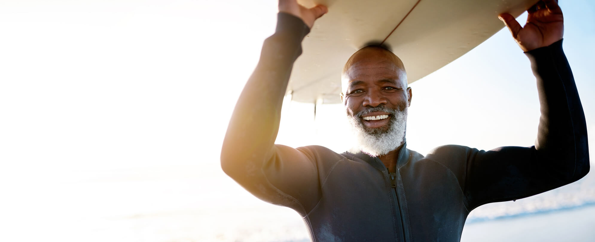 Older gentlemen in wetsuit smiling, carrying a surfboard on his head