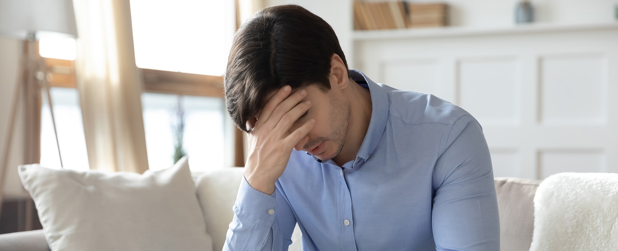 A frustrated man sitting on a couch puts his head in his hand.