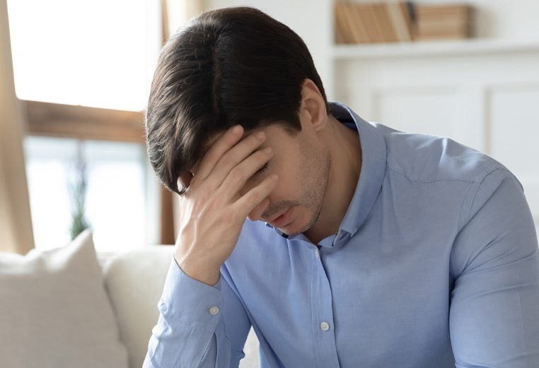 A frustrated man sitting on a couch puts his head in his hand.