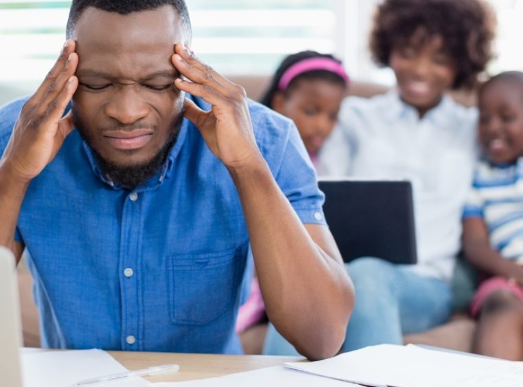 Stressed father sitting at the kitchen table, working on a laptop, holding his head with his wife and kids having fun in the background 