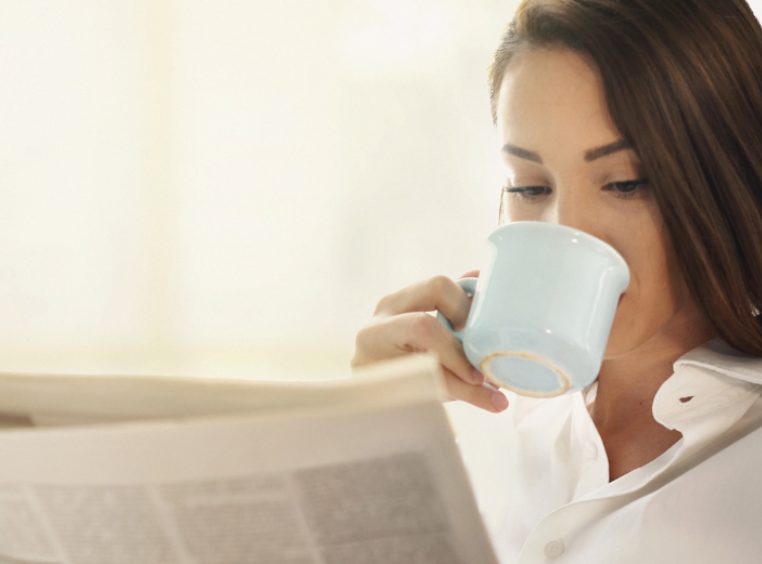 Woman sitting by a living room window, having a coffee and reading a newspapers. She has long brown hair and wearing a white shirt.