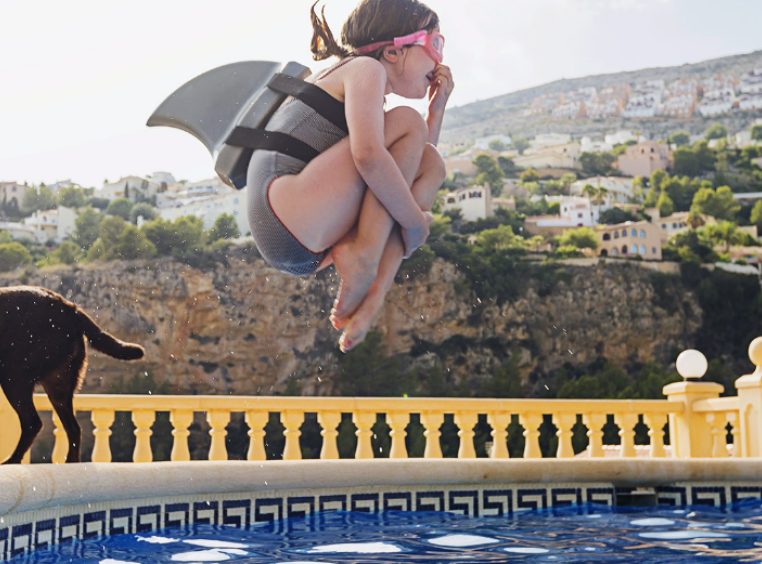 Young girl cannonballing into a pool with a shark fin float strapped to her back