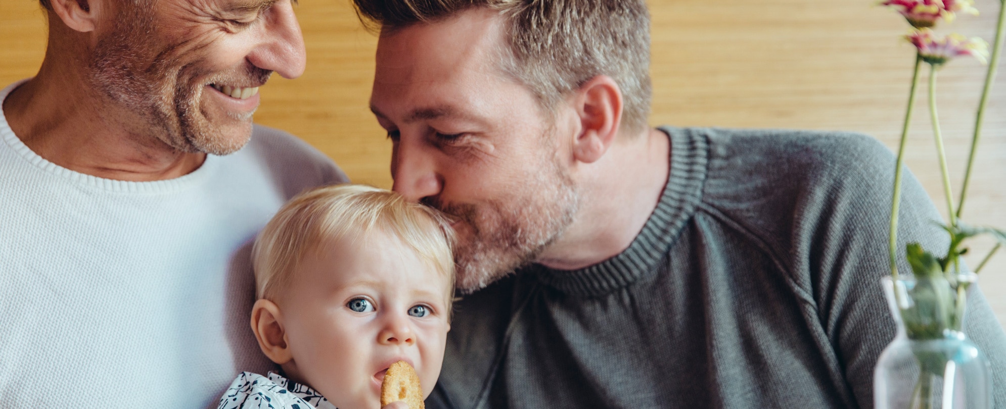 Happy gay couple holding their son. Father kisses son on the head as he eats his cookie. 