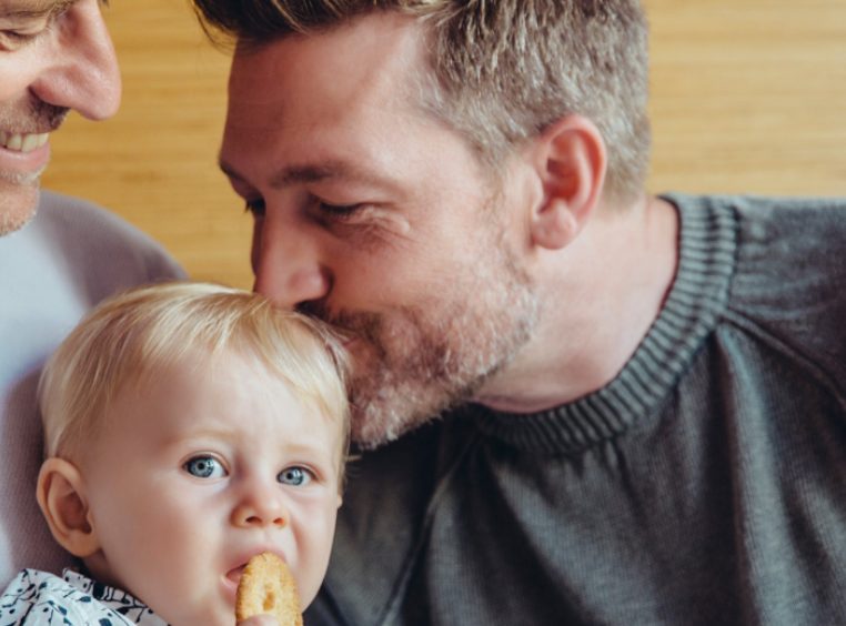 Happy gay couple holding their son. Father kisses son on the head as he eats his cookie. 
