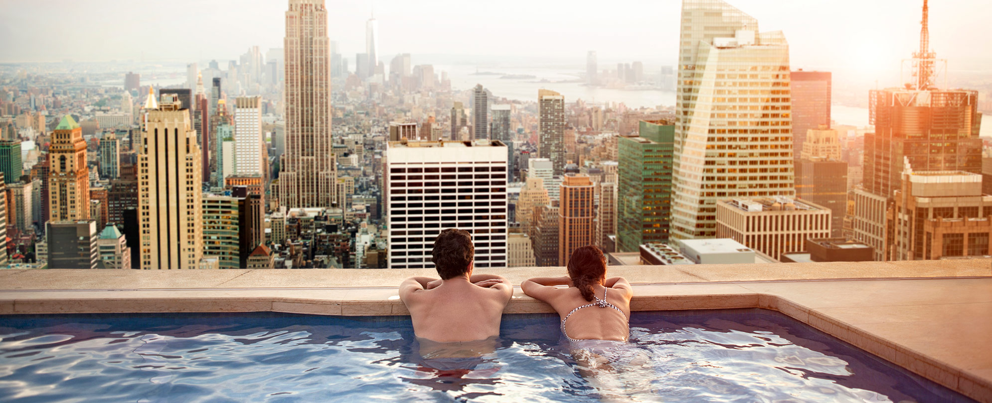 A man and a woman swimming in a rooftop pool overlooking the city skyline. 