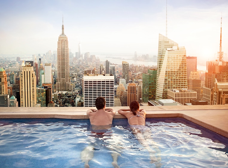 A man and a woman swimming in a rooftop pool overlooking the city skyline. 
