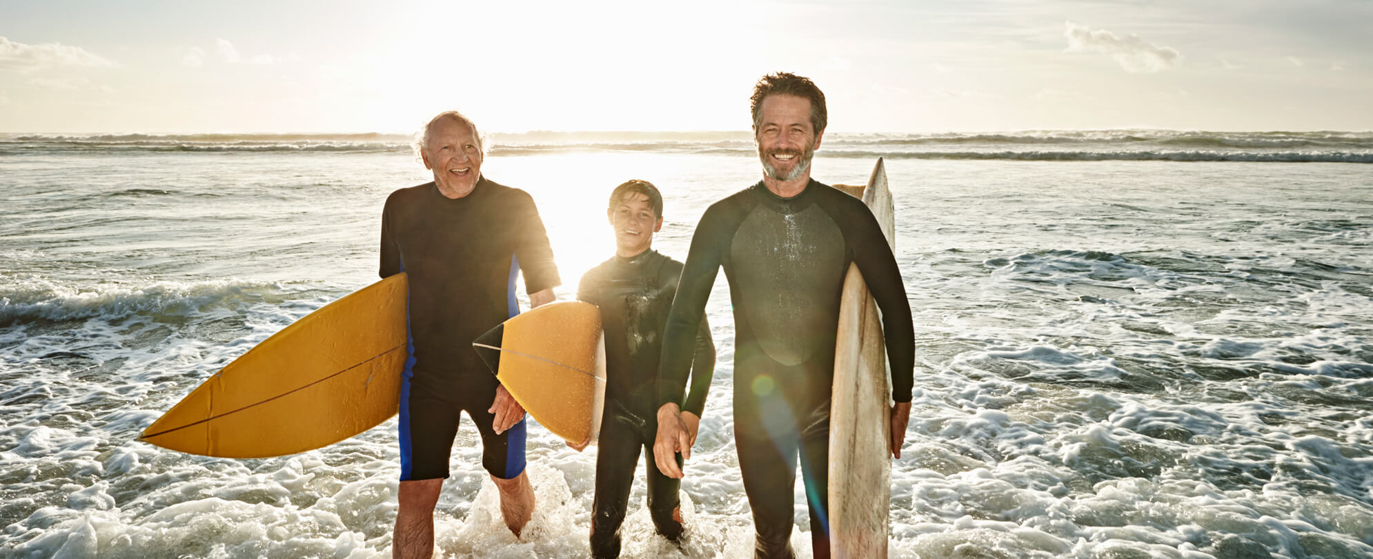 Grandfather, father, and son walking on to the shore with surfboards in hand 