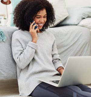 A woman sitting on the floor on her phone looking over documents on her laptop 