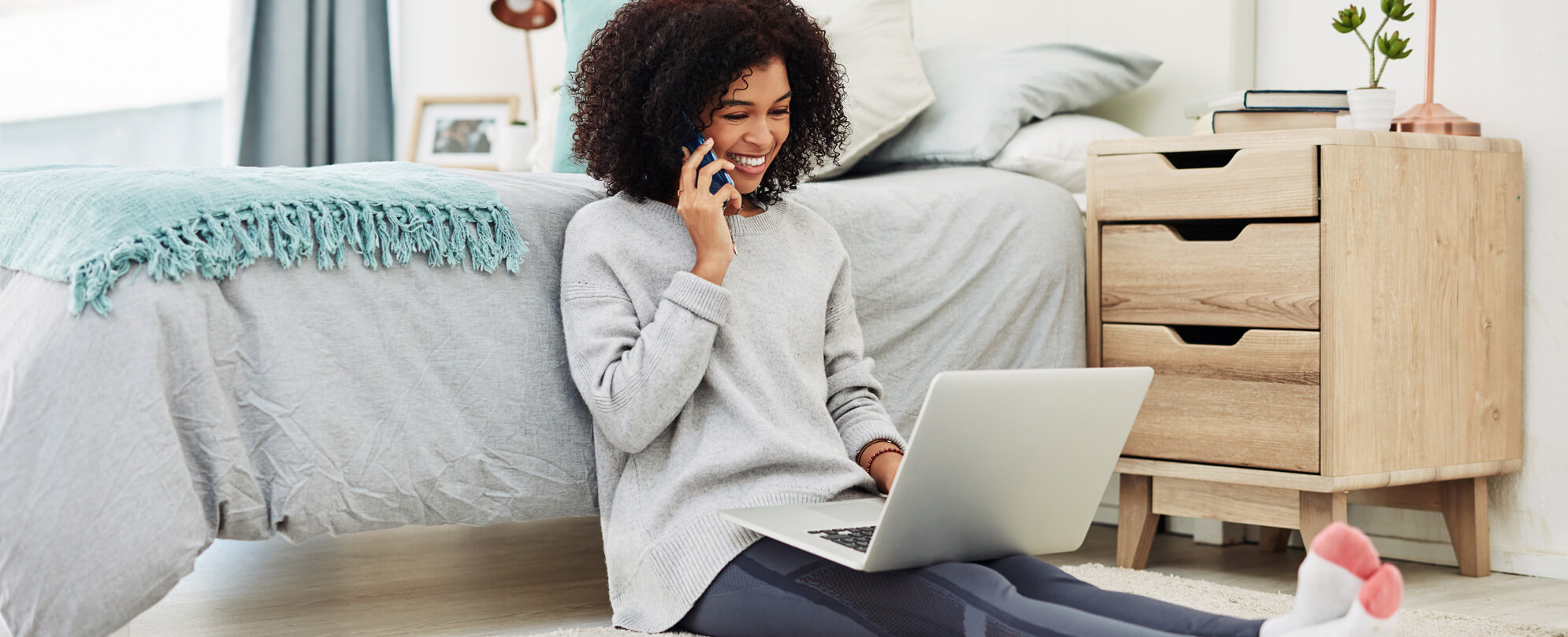 A woman sitting on the floor on her phone looking over documents on her laptop 